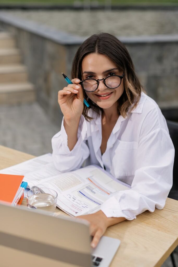 A young woman in glasses working remotely on her laptop with books and papers on a desk.